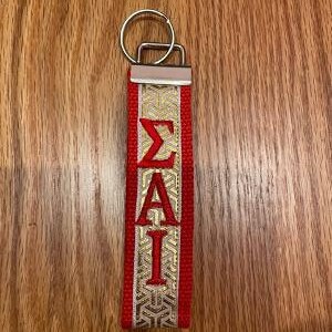 May include: Red and gold keychain with the Greek letters &Sigma;&Alpha;&Iota; embroidered in red. The keychain has a silver metal ring and a decorative gold pattern on the fabric.