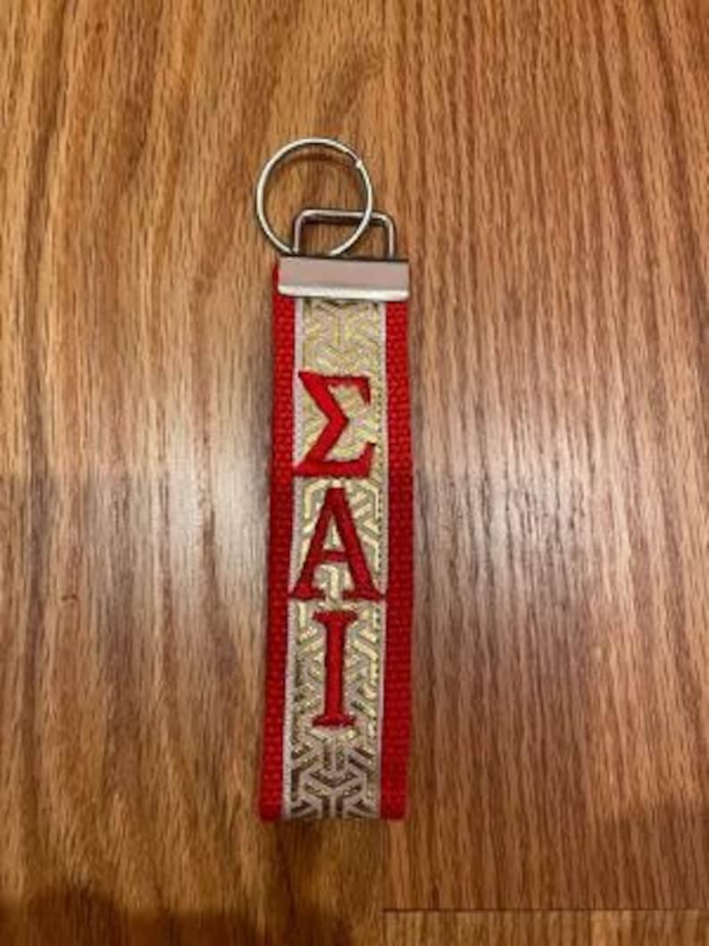 May include: Red and gold keychain with the Greek letters &Sigma;&Alpha;&Iota; embroidered in red. The keychain has a silver metal ring and a decorative gold pattern on the fabric.