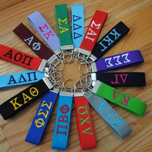 May include: A collection of 14 colorful keychains with embroidered Greek letters. The keychains are arranged in a circle on a wooden surface.