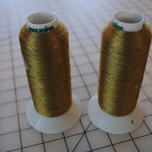 May include: Two spools of gold-colored Madeira brand thread. The thread is tightly wound on white spools. The spools are sitting on a white grid surface.