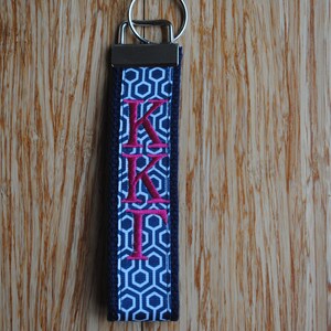 May include: A navy blue and white geometric pattern keychain wristlet with the embroidered letters KKI in pink. The keychain has a silver metal clasp and ring.