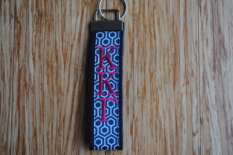 May include: A navy blue and white geometric pattern keychain wristlet with the embroidered letters KKI in pink. The keychain has a silver metal clasp and ring.