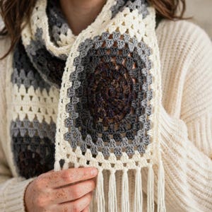 May include: A crochet scarf featuring a series of square motifs in a colour palette of cream, light grey, dark grey, and deep brown. The scarf is constructed with a classic granny square pattern and is finished with long cream-coloured fringe tassels.