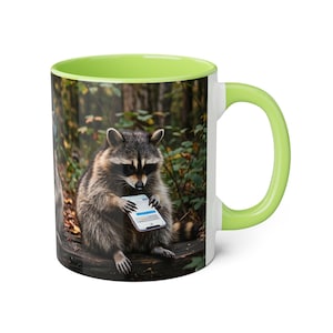 May include: White ceramic mug with a green interior and handle. The mug features a raccoon holding a smartphone, set against a forest backdrop. The raccoon is sitting on a log. The mug's design is printed on the exterior.