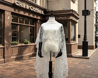 May include: A white lace shawl with a scalloped edge is displayed on a mannequin. The shawl is draped over the mannequin's shoulders and extends down to the ground. The background includes a cafe with the text "CAFE Coffee &" on the building.