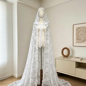 May include: A white lace hooded cape displayed on a mannequin. The cape features intricate floral patterns and a long train. The delicate fabric drapes gracefully, showcasing the cape's design. The cape is a bridal or formal wear item.