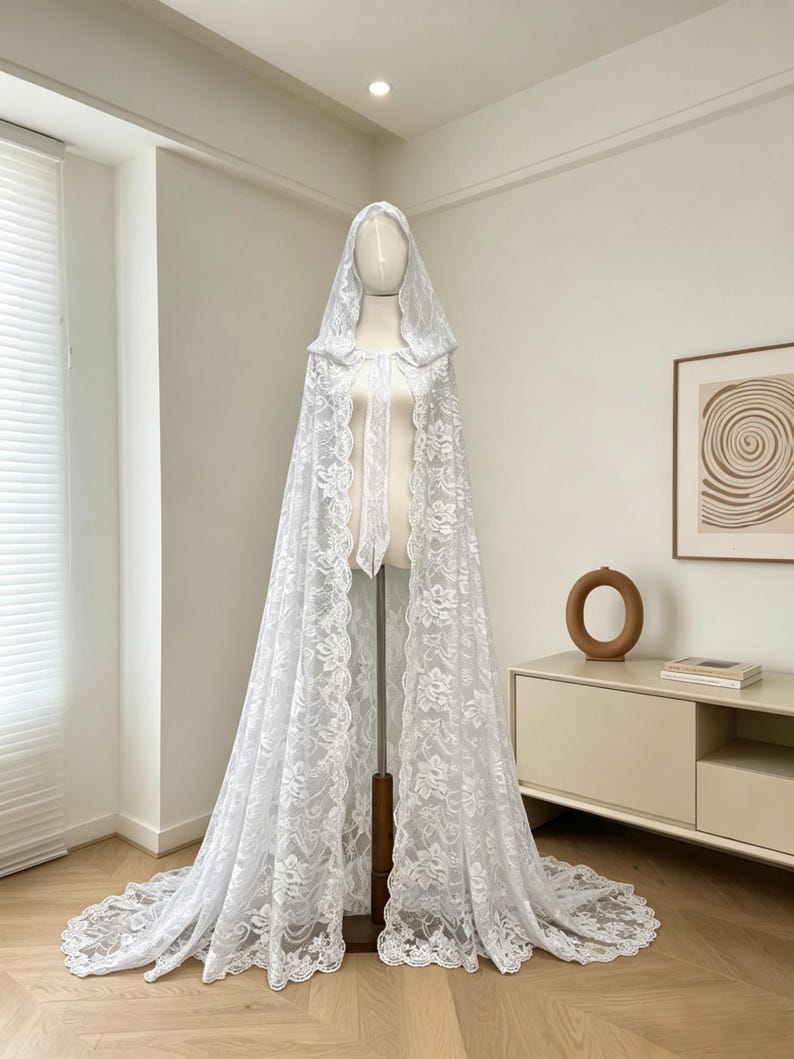 May include: A white lace hooded cape displayed on a mannequin. The cape features intricate floral patterns and a long train. The delicate fabric drapes gracefully, showcasing the cape's design. The cape is a bridal or formal wear item.