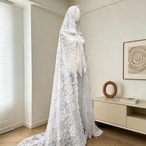 May include: A white lace hooded cape with a long train. The cape is displayed on a mannequin, showcasing the intricate floral lace design. The cape features a tie closure at the neck and a scalloped edge.