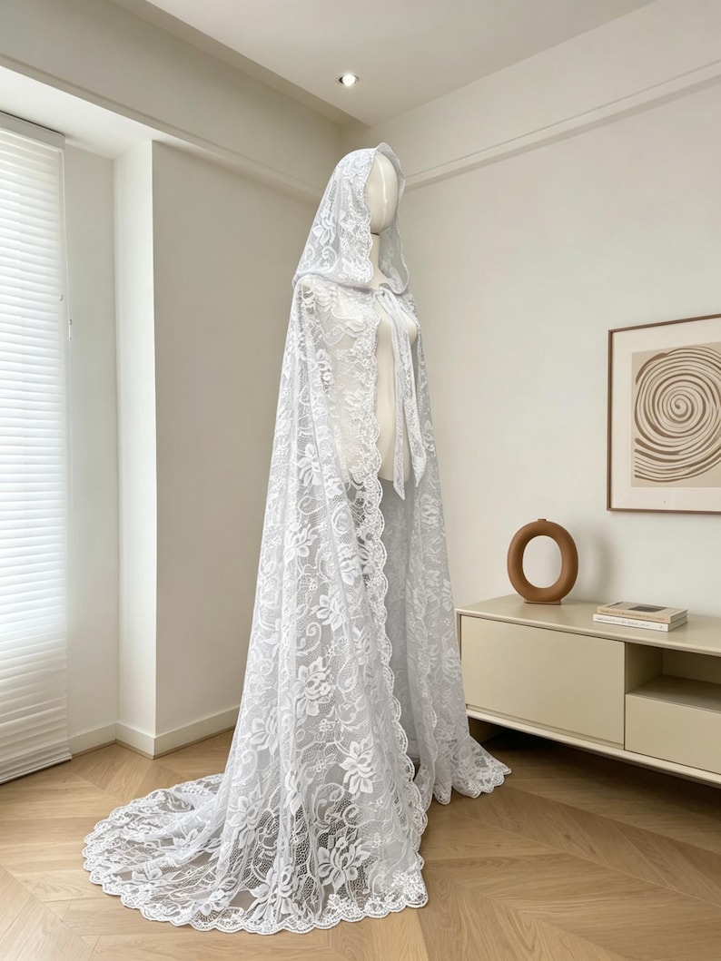 May include: A white lace hooded cape with a long train. The cape is displayed on a mannequin, showcasing the intricate floral lace design. The cape features a tie closure at the neck and a scalloped edge.