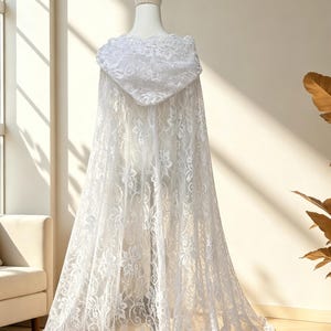 May include: A white lace hooded cape displayed on a mannequin. The cape features a floral pattern and a long, flowing design. The hood is also adorned with lace, creating a delicate and elegant look. The cape is suitable for weddings or special occasions.