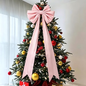 Op de afbeelding: Een versierde kerstboom met een grote, lichtroze stoffen strik aan de top, gouden en rode kerstballen, warmwitte kerstverlichting en een gouden ster als piek. Rode en gouden cadeaus staan aan de voet van de boom.