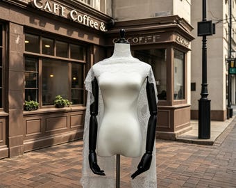 May include: A white dress form with black arms and a black head post displays a white lace shawl. The dress form is on a brick-paved street in front of a cafe with the words "CAFE Coffee &" on the building.