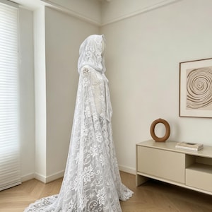 May include: A white lace hooded cape with a floral pattern. The cape has a long train and is displayed on a mannequin. The lace is detailed and the cape appears to be made of a delicate fabric.