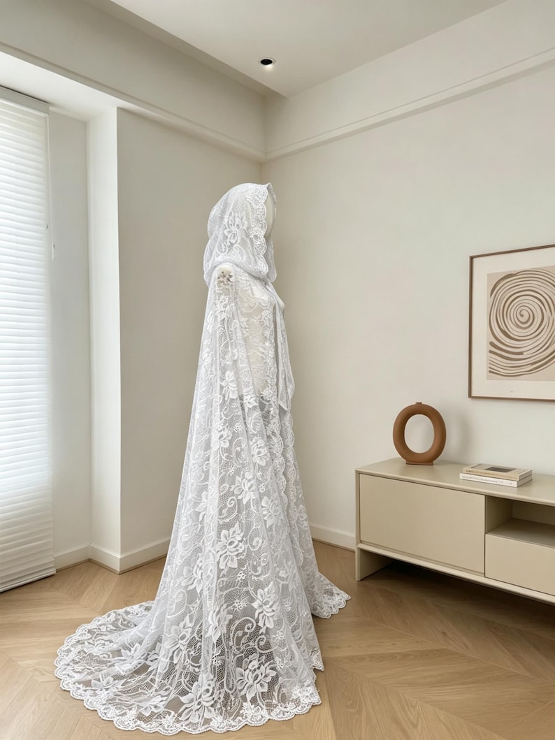 May include: A white lace hooded cape with a floral pattern. The cape has a long train and is displayed on a mannequin. The lace is detailed and the cape appears to be made of a delicate fabric.