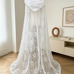 May include: A white lace hooded cape with floral patterns. The cape is displayed on a mannequin, showcasing its full length and intricate design. The lace fabric drapes elegantly, creating a flowing silhouette. The hood is also adorned with lace.