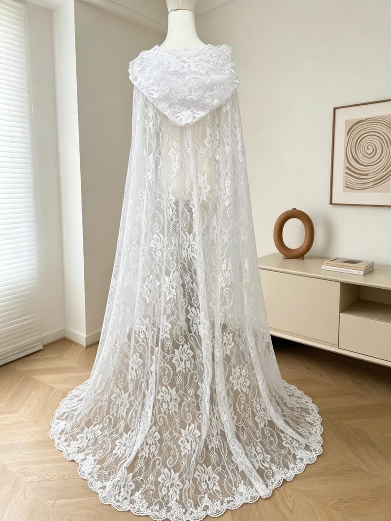 May include: A white lace hooded cape with floral patterns. The cape is displayed on a mannequin, showcasing its full length and intricate design. The lace fabric drapes elegantly, creating a flowing silhouette. The hood is also adorned with lace.