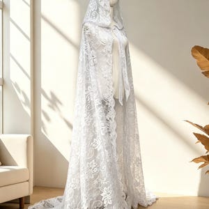 May include: A white lace hooded cape with floral patterns. The cape is displayed on a mannequin, showcasing its flowing design and long train. The intricate lace detailing adds a delicate and elegant touch to the garment.