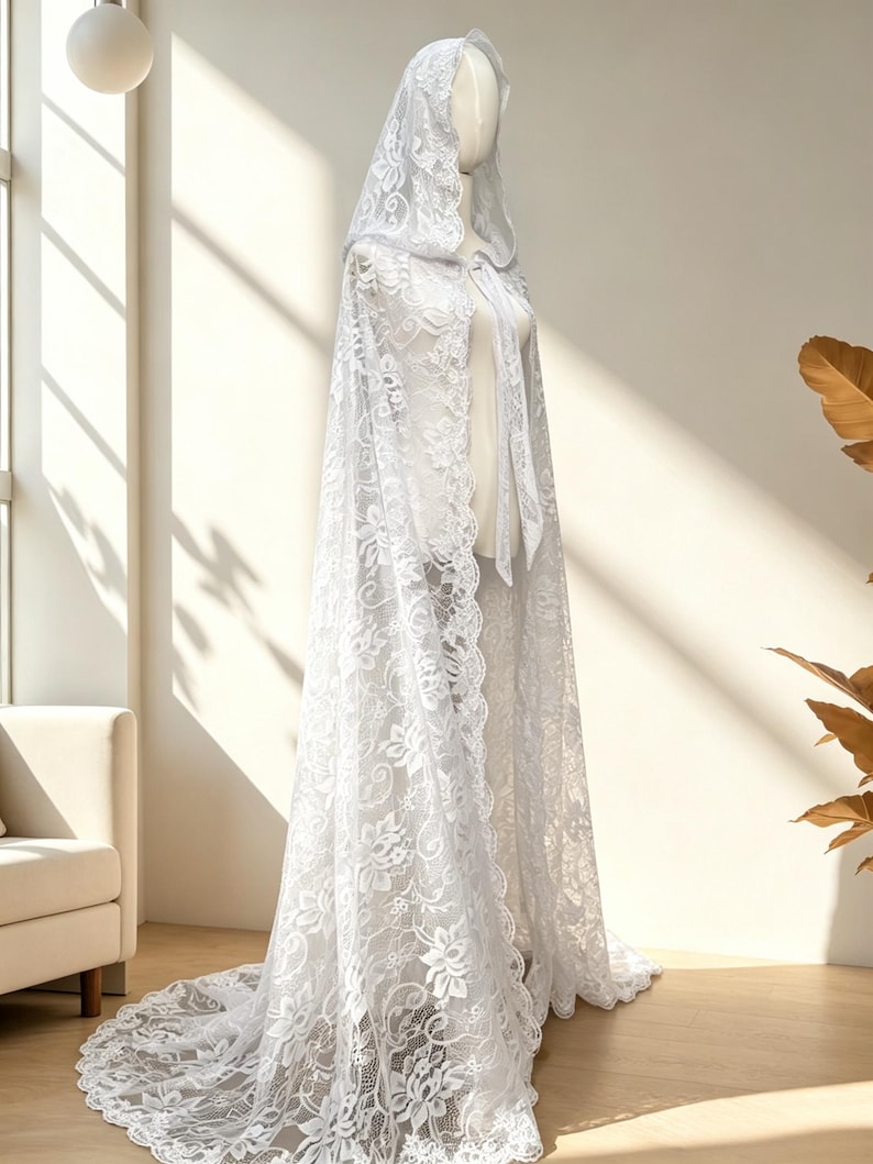 May include: A white lace hooded cape with floral patterns. The cape is displayed on a mannequin, showcasing its flowing design and long train. The intricate lace detailing adds a delicate and elegant touch to the garment.