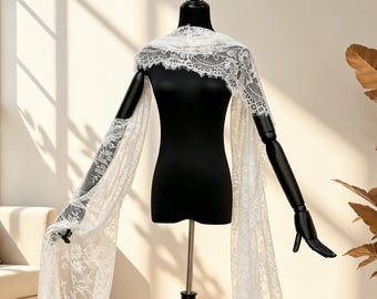 May include: A white lace shawl with long sleeves is draped over a black mannequin. The delicate lace features intricate floral patterns. The mannequin is on a black stand, and the background is a neutral color.