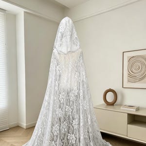 May include: A white lace hooded cape, displayed on a mannequin. The cape features a floral pattern and a long, flowing design. The hood is also made of the same delicate lace material.