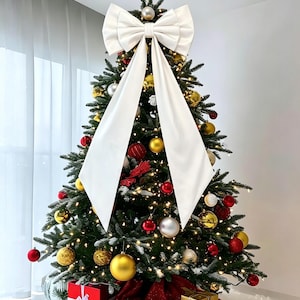 May include: A large white fabric bow topper for a Christmas tree, featuring long trailing ribbons. The tree is decorated with gold and red spherical ornaments, warm white string lights, and a gold star at the very top, with gifts below.