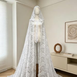 May include: A white lace hooded cape displayed on a mannequin. The cape features a floral lace pattern and a long train. The mannequin is on a wooden stand, and the background is a neutral-toned room with a cabinet and artwork.