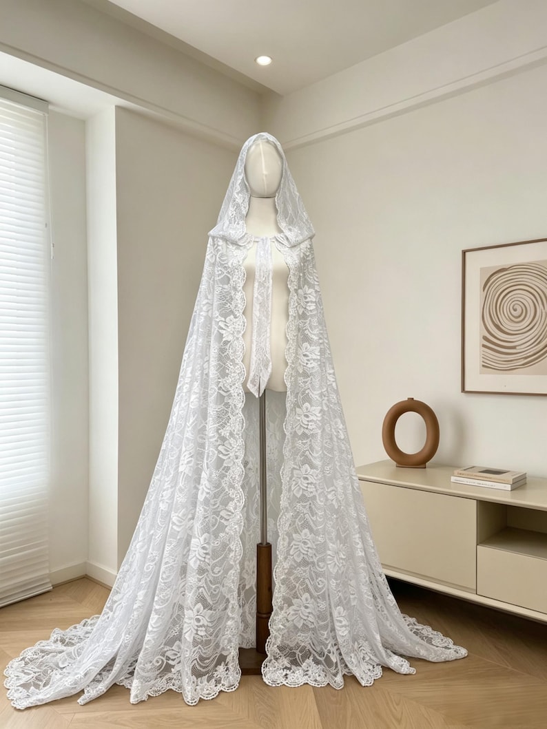 May include: A white lace hooded cape displayed on a mannequin. The cape features a floral lace pattern and a long train. The mannequin is on a wooden stand, and the background is a neutral-toned room with a cabinet and artwork.