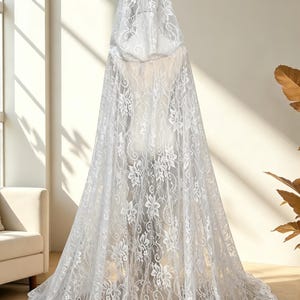 May include: A white lace hooded cloak with a floral pattern. The cloak is long, flowing, and covers the entire form. The lace is detailed and delicate, creating a romantic and ethereal look. The cloak is displayed on a mannequin.