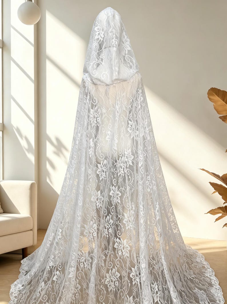 May include: A white lace hooded cloak with a floral pattern. The cloak is long, flowing, and covers the entire form. The lace is detailed and delicate, creating a romantic and ethereal look. The cloak is displayed on a mannequin.
