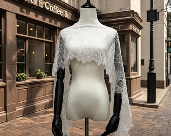 May include: A white lace shawl draped over a mannequin with black arms. The shawl features an intricate floral pattern with scalloped edges that extend down the sides. The fabric is sheer and lightweight, covering the shoulders and upper torso.