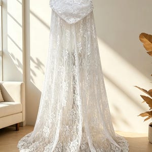May include: A white lace hooded cape displayed on a mannequin. The cape features a floral lace pattern and a flowing design. The hood and hemline are intricately detailed. The cape is a bridal or formal wear item.