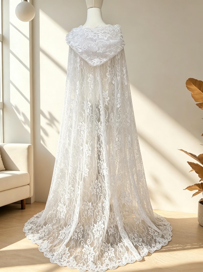 May include: A white lace hooded cape displayed on a mannequin. The cape features a floral lace pattern and a flowing design. The hood and hemline are intricately detailed. The cape is a bridal or formal wear item.