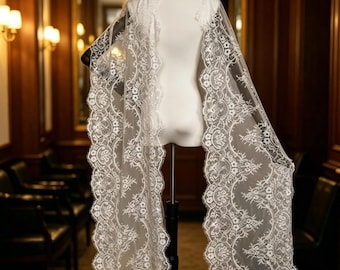 May include: A long, white lace shawl featuring an intricate floral pattern with scalloped edges. The sheer fabric is draped over a mannequin, showcasing the detailed embroidery and delicate mesh construction of the textile accessory.