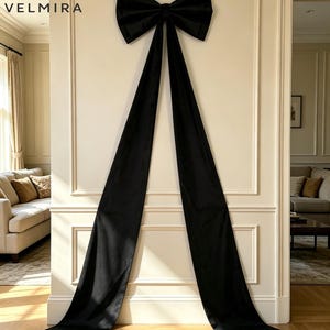 May include: A large black decorative bow with two long, trailing fabric ribbons hanging vertically. The bow is centered on a wall panel. The text VELMIRA with a crescent moon and feather icon is visible in the upper left corner of the image.