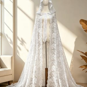 May include: A white lace hooded cape displayed on a mannequin. The cape features intricate floral patterns and a long train. The fabric appears delicate and flowing, with a soft, romantic aesthetic. The cape is designed for special occasions.