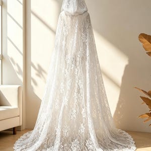 May include: A white lace hooded cape with a long train. The cape is made of delicate floral lace and drapes to the floor. The hood is also lace, and the cape is displayed in a well-lit room.