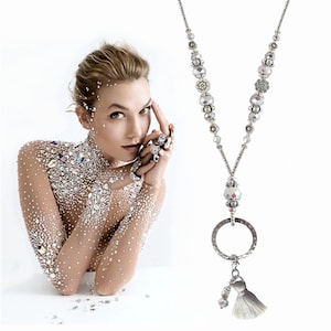 May include: A silver beaded necklace with a silver ring and a grey tassel. The necklace features a variety of silver beads, including round beads, flower-shaped beads, and faceted beads.