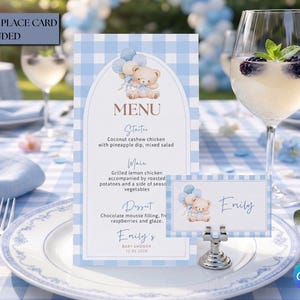 May include: A baby shower table setting with a blue and white gingham tablecloth. The setting includes a menu card, place card, and a wine glass with a drink and berries. A napkin ring shaped like a teddy bear is also present.