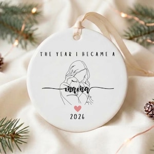 Op de afbeelding: Een rond wit keramieken ornament met een zwarte lijntekening van een persoon die een baby vasthoudt. De tekst luidt THE YEAR I BECAME A mama met een klein roze hartje en het jaartal 2026 eronder gedrukt. Een crèmekleurig lint is bovenaan bevestigd.