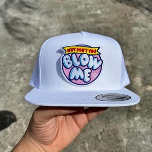 Op de afbeelding: Witte truckerpet met een mesh achterkant en een platte klep. De pet heeft een patch met de tekst "WHY DON'T YOU BLOW ME" in een bubble letter stijl, met roze en blauwe accenten.