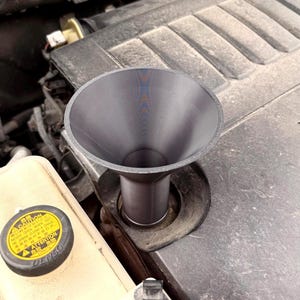 May include: A dark gray plastic funnel is positioned over an engine component. The engine bay includes a coolant reservoir with a yellow cap and the text "Caution". The engine block has a "V6" emblem.