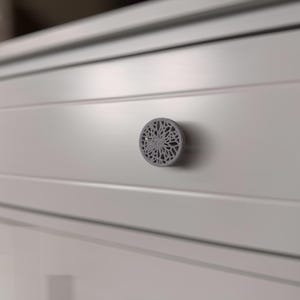 May include: A close-up of a white cabinet drawer with a gray, circular decorative knob. The knob features an intricate, openwork floral design. The cabinet has a simple, modern design.