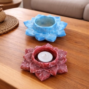 May include: Two decorative lotus flower candle holders. One is blue, the other is red with a lit white candle inside. The holders are made of a textured material and sit on a wooden table. Home decor.