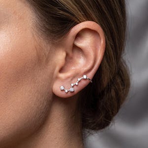 May include: A silver-toned ear crawler earring featuring a constellation-inspired design with multiple small, clear, round-cut stones set along a curved metal bar that follows the natural shape of the earlobe and cartilage.