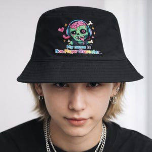 Könnte beinhalten: Schwarzer Fischerhut mit einem farbenfrohen Cartoon-Zombie-Gehirn, das Kopfhörer trägt. Der Zombie hat grüne Haut und streckt die Zunge heraus. Auf dem Hut steht der Text "My name is Non-Player Character..."