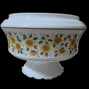May include: A white ceramic pedestal bowl featuring a continuous wrap-around pattern of yellow sunflowers with green leaves and grey stems. The bowl has a wide, flared rim and a scalloped base, accented by thin brown lines along the top and bottom.