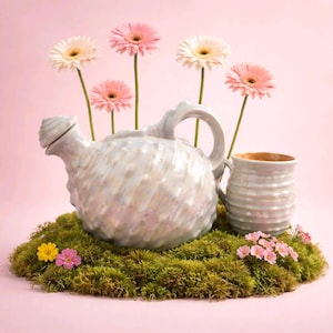 Può includere: Un set di annaffiatoio e tazza in ceramica blu chiaro con smalto iridescente, appoggiato su muschio verde. Margherite gerbera rosa e bianche sono disposte dietro l'annaffiatoio. Lo sfondo è rosa chiaro.