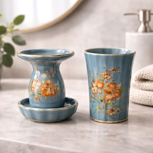 Pode incluir: Um conjunto de saboneteira e copo de cerâmica azul. O conjunto apresenta um design floral com flores laranja e amarelas e folhas verdes. As bordas são aparadas com dourado. A saboneteira tem uma base de pedestal.