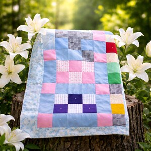 Może przedstawiać: Pikowana kołdra patchwork z niebieską kwiatową obwódką, z kwadratami z różowych, niebieskich i wzorzystych tkanin. Kołdra jest prezentowana na zewnątrz, spoczywając na pniu drzewa, z białymi liliami w tle.