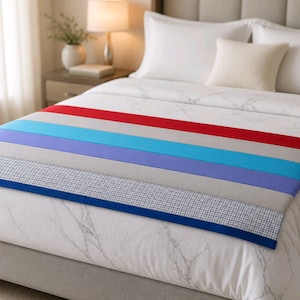 May include: A colorful bed runner with horizontal stripes of red, light blue, purple, and gray. The bottom edge features a blue border and a checkered pattern. The bed runner is placed on a white bed with pillows.